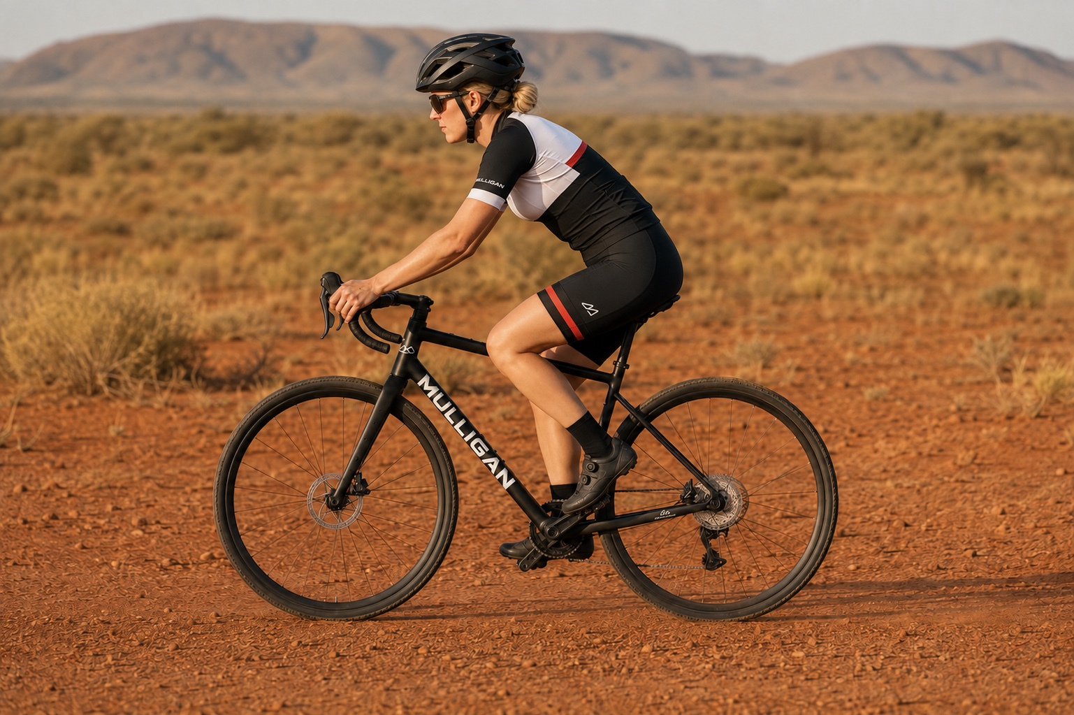 Vélo gravel aluminium G2 MULLIGAN noir sur une piste de terre rouge avec une cycliste roulant de droite à gauche de profil.
